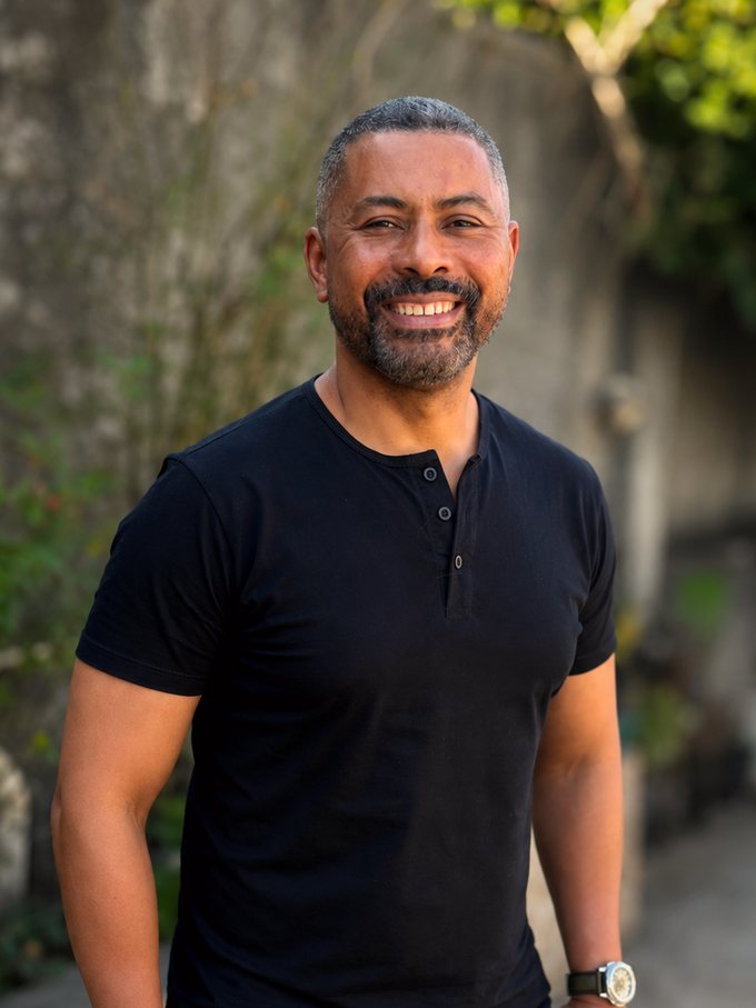 Ademir F. Silva, de camiseta preta, sorrindo em uma viela arborizada.